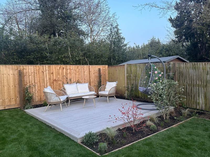 A pale grey deck with outdoor sofa and chairs, an edged planting bed and new turf, closeboard fencing behind