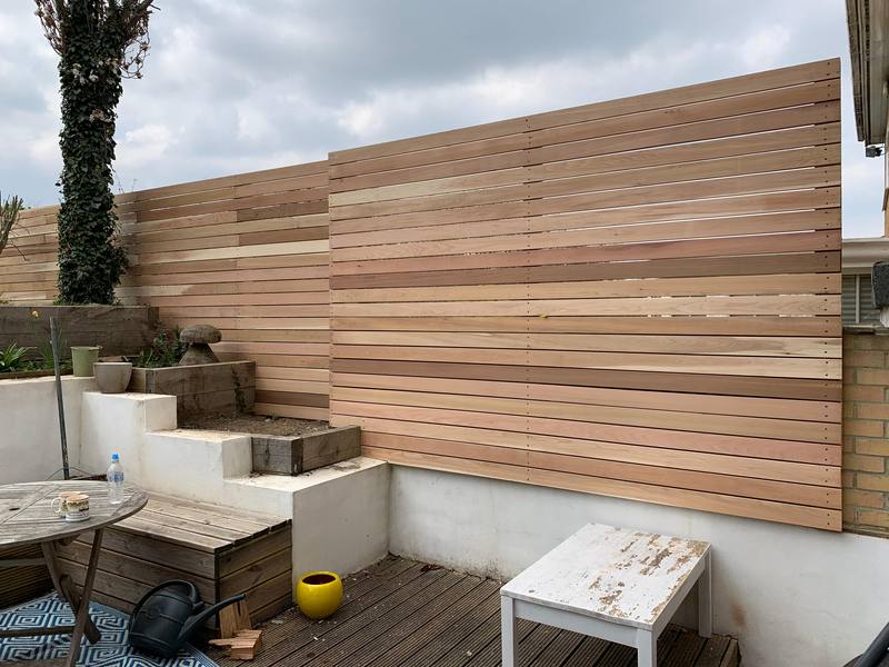 A tall horizontal slatted timber screen above white rendered raised beds and decking