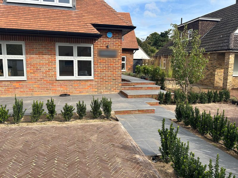 Young hedging plants set out along a new grey paved path and steps, herringbone block paving in front of a brick house