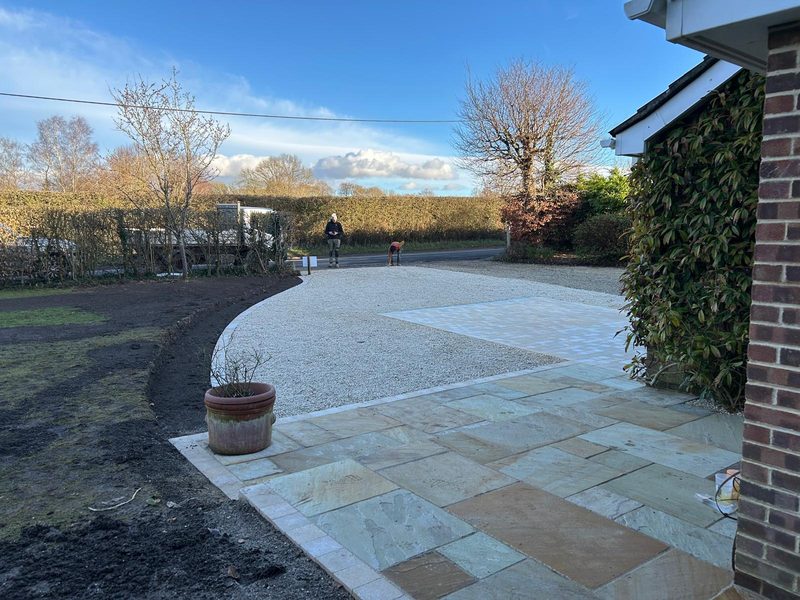 A stone patio in mixed buff and grey tones with a pale block edge, opening onto a curved gravel area beside a brick house