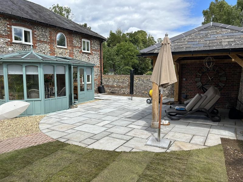 A newly laid stone patio with a curved edge, between a flint-and-brick house with a conservatory and an open-sided garden building, new turf in the foreground