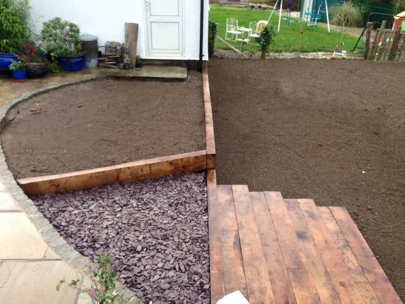Sleeper-edged beds of freshly prepared soil beside slate chippings and a flight of timber sleeper steps, a stone patio with a sett edge in the corner