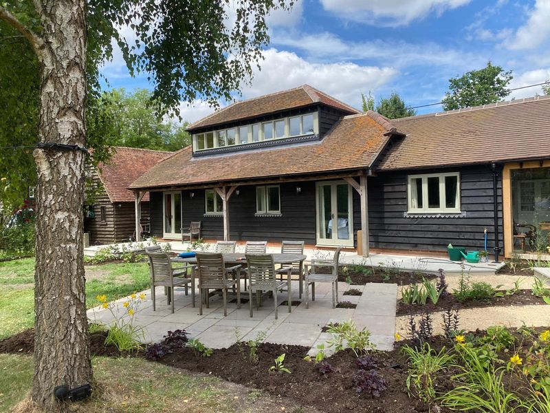 The barn's stone patio and dining set seen past a silver birch trunk, young plants in the new borders