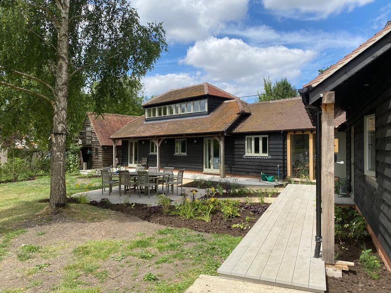 Wide view of the barn conversion: a stone patio with a dining table, a new lawn and freshly planted borders, a walkway along the oak-framed colonnade