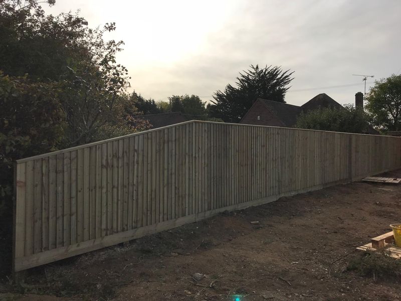 A long new closeboard fence with gravel boards along its foot, running beside bare, cleared ground, houses and trees behind