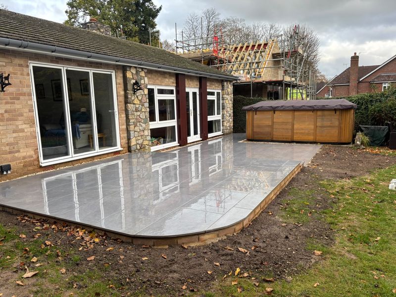 The curved end of a large-format grey patio on a buff-brick edge, wet after rain, a timber-clad hot tub at the far side