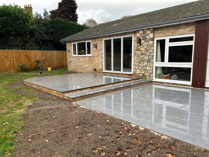 Large-format grey paving on two levels behind a bungalow, with buff-brick risers and a gravel strip along the house
