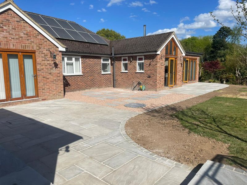A newly laid stone patio with a curved, sett-edged border behind a brick bungalow, bare soil beside it
