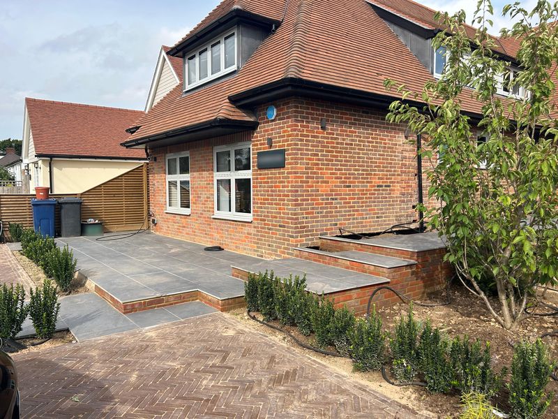The front of a brick house: grey paving on brick-built steps and a raised bed, herringbone block paving, and a row of young hedging plants
