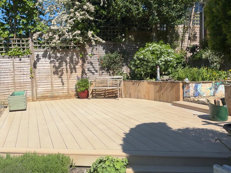 A large pale-boarded deck with a step down to the garden, a timber-clad raised bed and a mosaic panel behind it, a bench and planters on the deck and lattice-topped fencing beyond