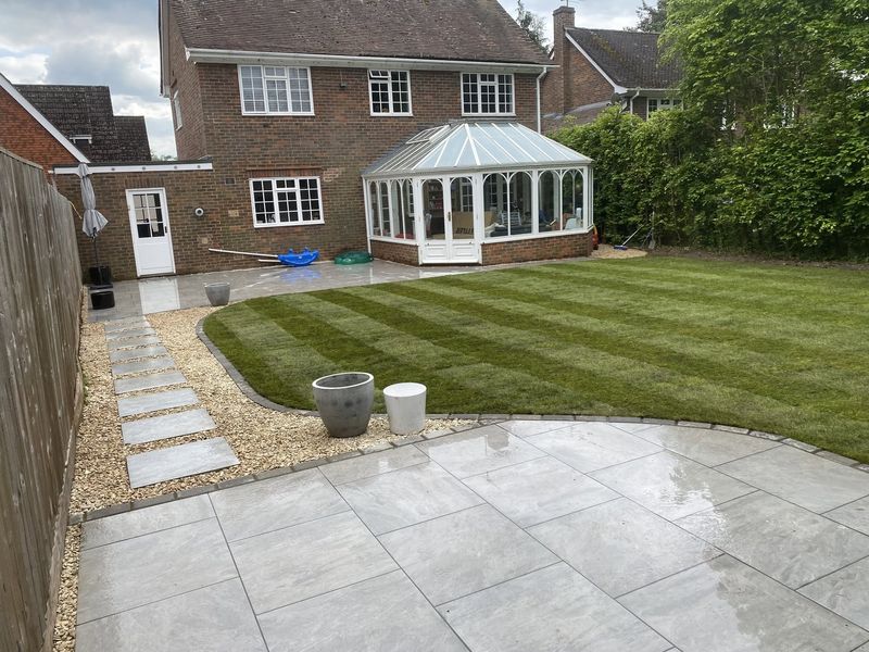 A grey paved patio behind a brick house with a conservatory, a striped lawn with a curved edge and a stepping-stone path through gravel