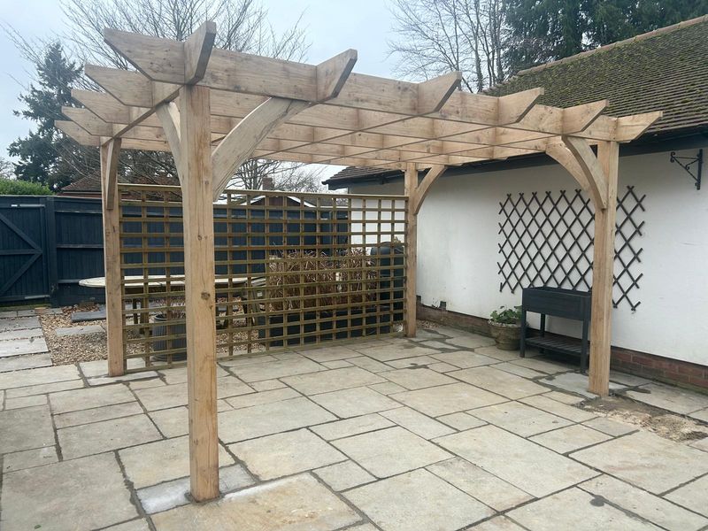 A timber pergola with a square trellis panel standing over a stone patio beside a white-rendered wall