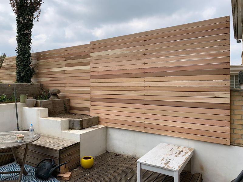 A tall horizontal slatted screen fixed above white rendered walls and raised beds, a deck and garden table in front