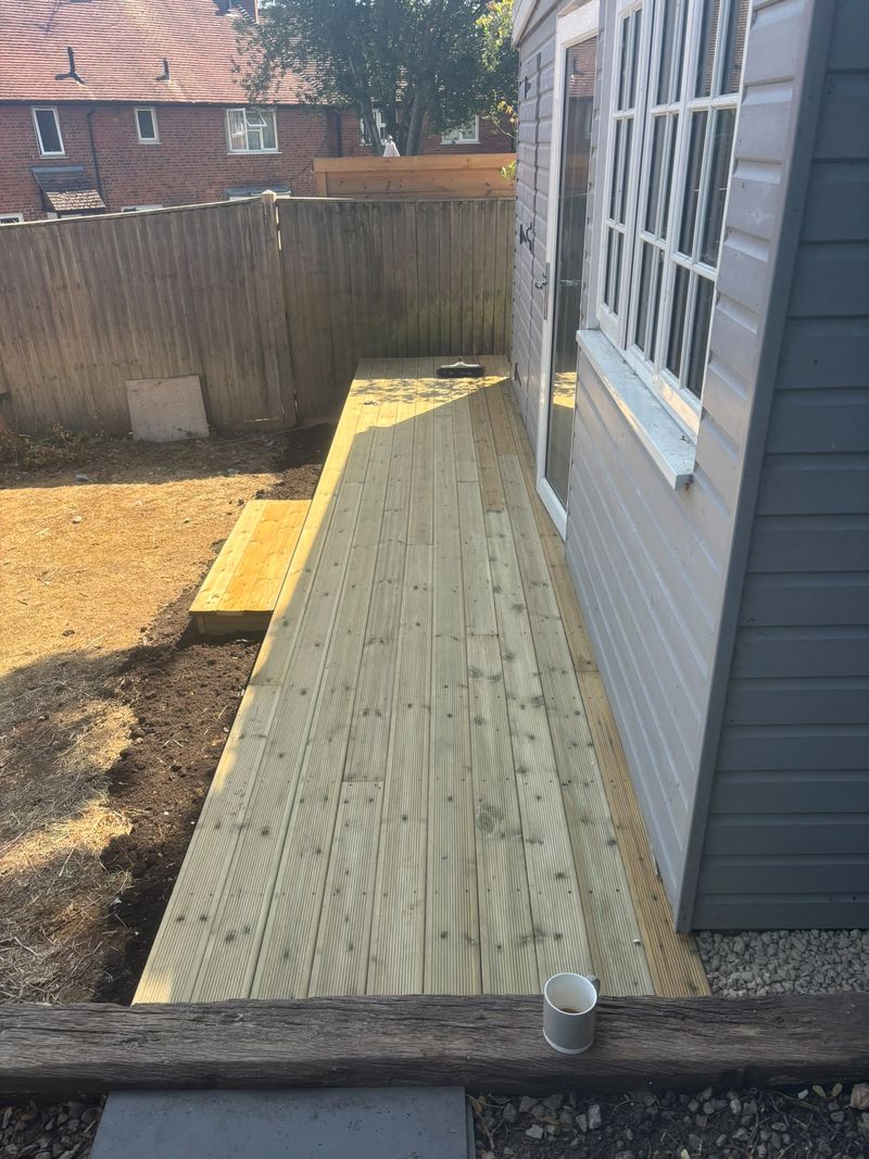 Looking along the new deck past the summerhouse doors and window to a closeboard fence, a box step at the side and an old sleeper across the foreground