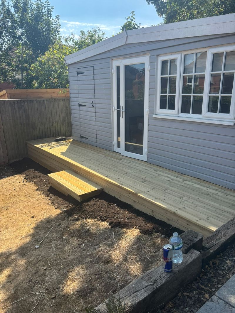 The new deck in front of the summerhouse's glazed doors, with boarded sides, a box step down to fresh soil and old sleepers in the foreground