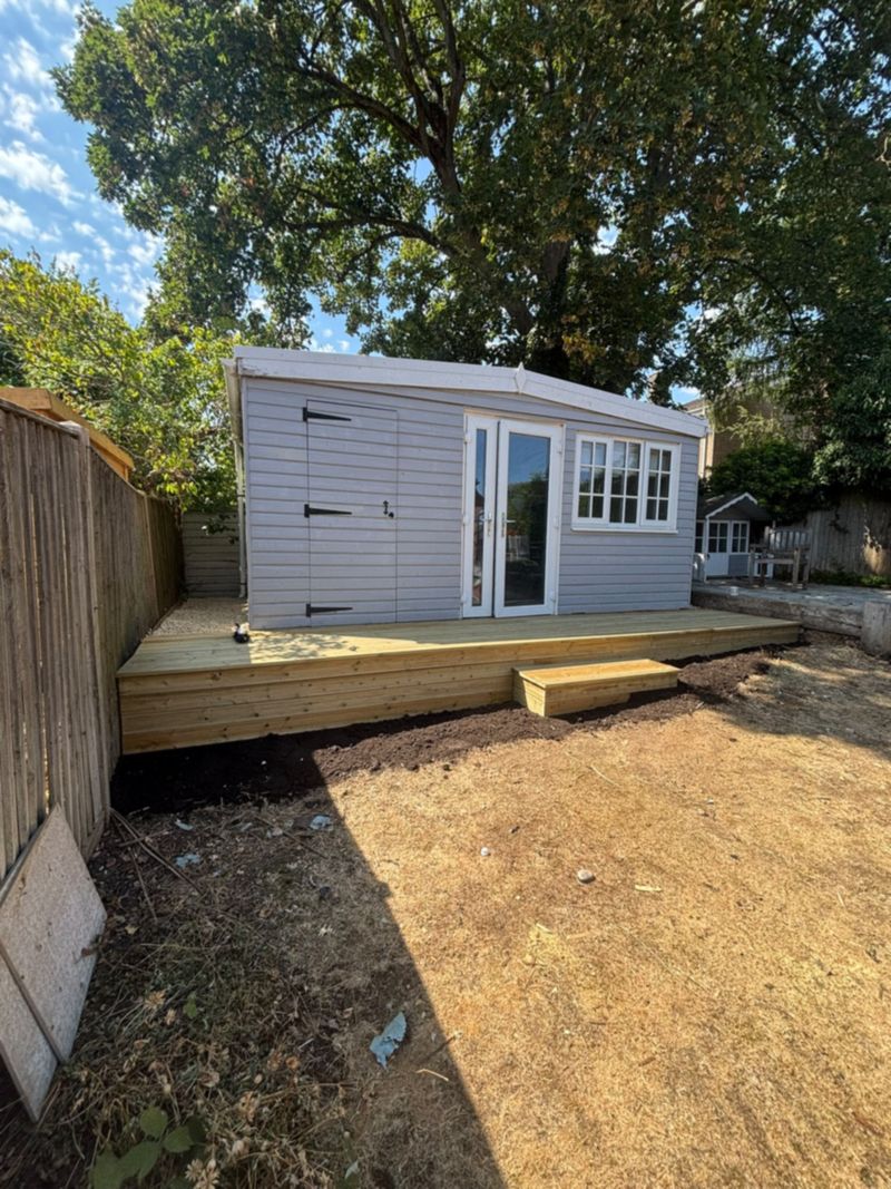 A new deck running across the front of a grey summerhouse with white doors and windows, a box step down to bare soil and dry grass, a large tree overhead