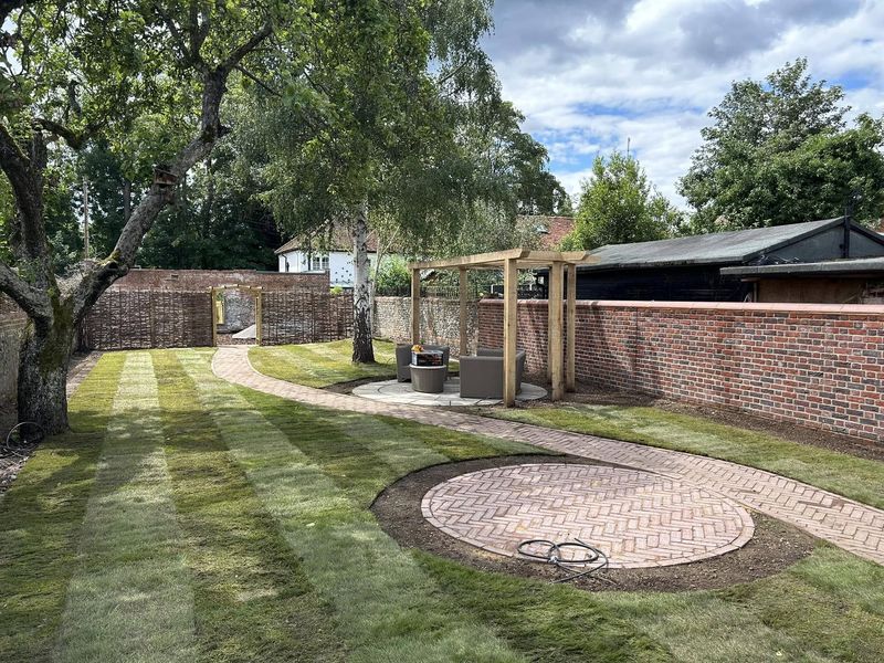 A finished walled garden: striped new turf, a curving herringbone brick path, a circular brick feature and a timber pergola over a round stone patio
