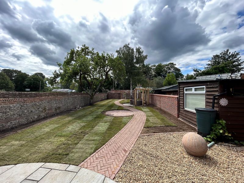 After: striped new turf either side of a curving herringbone brick path, gravel and a stone ball in the foreground, a pergola at the far end