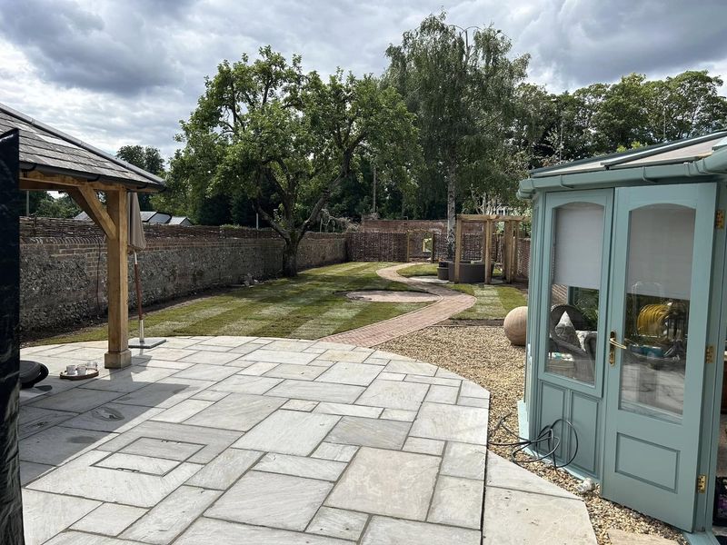A natural stone patio laid in mixed sizes between a timber-framed gazebo and a garden room, with the new lawn and brick path beyond