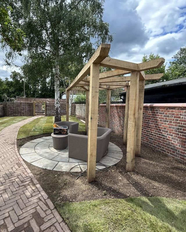A timber pergola standing over a circular stone patio with garden seating, next to a red-brick wall, the herringbone path curving past